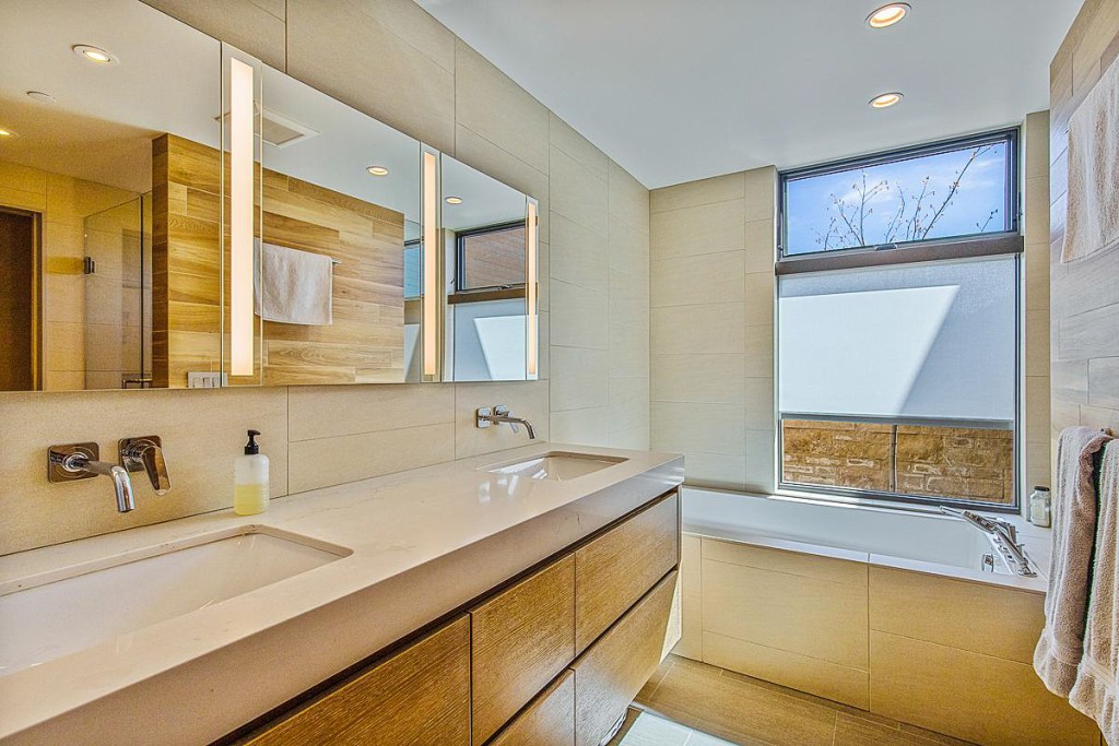 Modern bathroom featuring a double sink vanity with wooden cabinetry, bright natural light from the window, and minimalist design elements.
