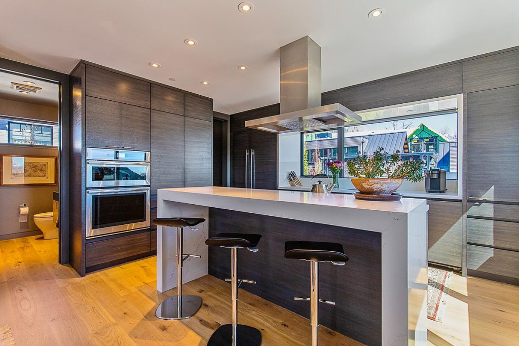 Modern kitchen featuring a sleek design with dark cabinetry, a white countertop, and bar stools, illuminated by natural light from large windows.
