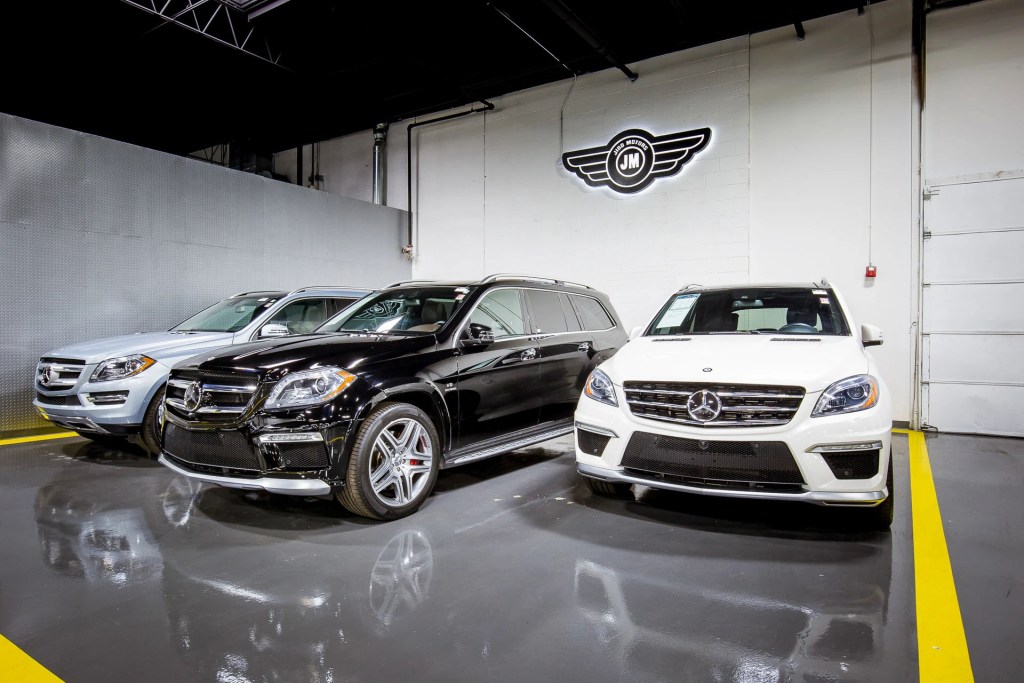 Three luxury vehicles, including a black SUV, a silver SUV, and a white SUV, are showcased in a well-lit car dealership. The dealership's logo is visible on the wall behind the cars.
