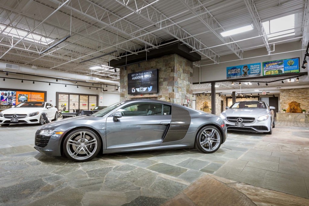 Interior view of a luxury car dealership showcasing a silver sports car in the foreground and a white luxury vehicle in the background, with modern decor and advertising displays.