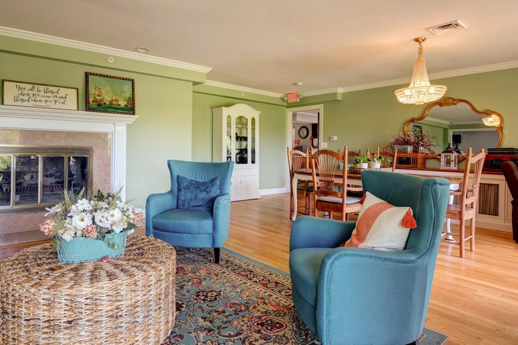 Cozy interior of a senior living space featuring two blue armchairs, a woven coffee table, and a dining area with wooden furniture and green walls.