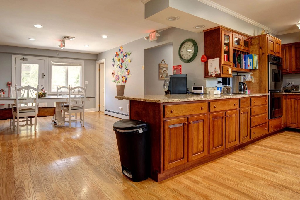 Interior view of a cozy kitchen area in a senior living community, featuring wooden cabinets, a dining table with chairs, and a colorful flower mural on the wall.