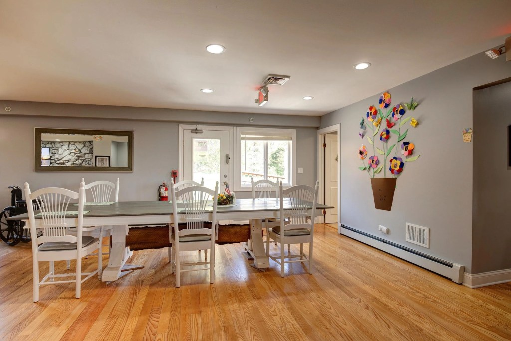 Interior view of a senior living community dining area featuring a large table with white chairs, decorated with a wall mural of a flower pot and colorful flowers.