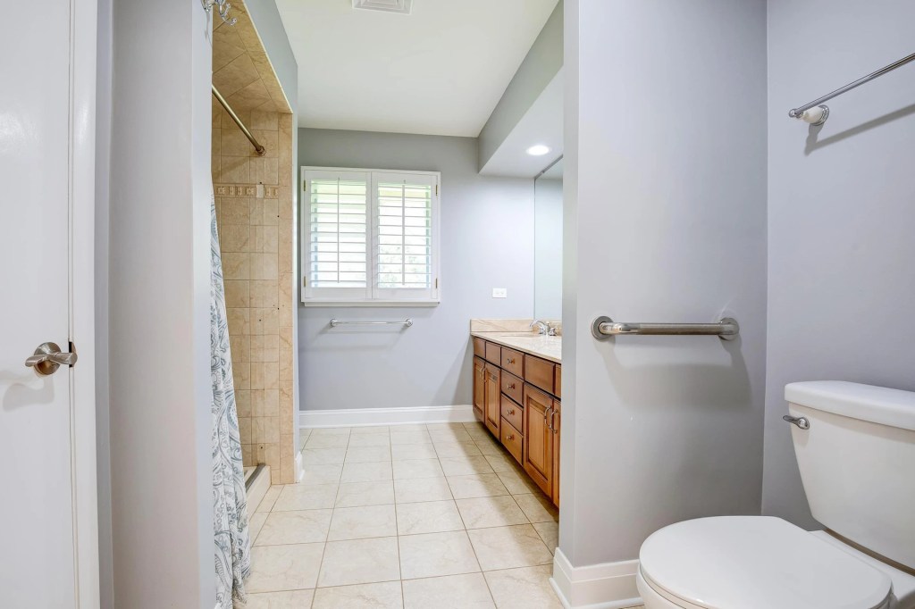 A modern bathroom with a walk-in shower, wooden vanity, and tiled floor. Natural light comes through a window with shutters.