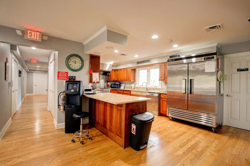 Interior view of a kitchen in a senior living community, featuring wooden cabinets, a granite countertop, stainless steel appliances, and a view of the hallway.