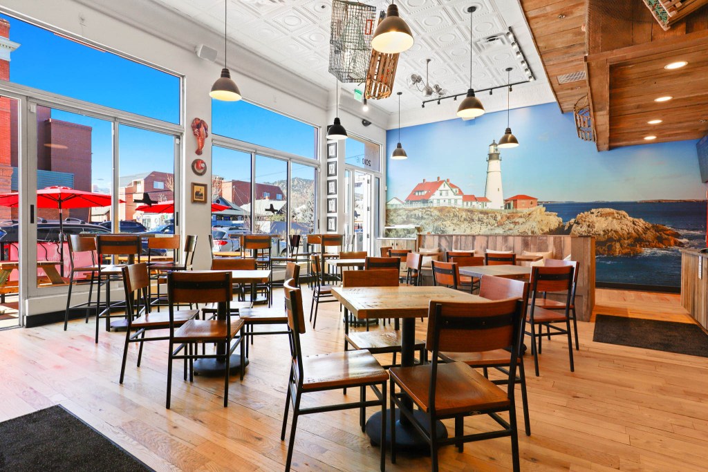 Interior view of a restaurant featuring wooden tables and chairs, large windows with outdoor seating visible, and a scenic mural of a lighthouse on the back wall.