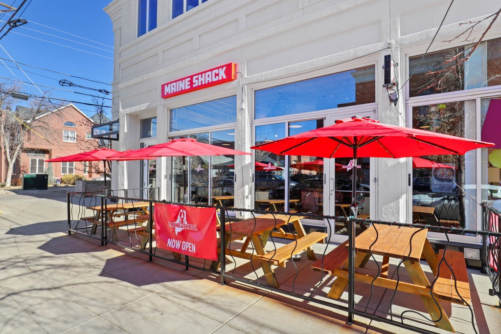 Exterior view of Maine Shack restaurant with red umbrellas over picnic tables and 'NOW OPEN' sign.