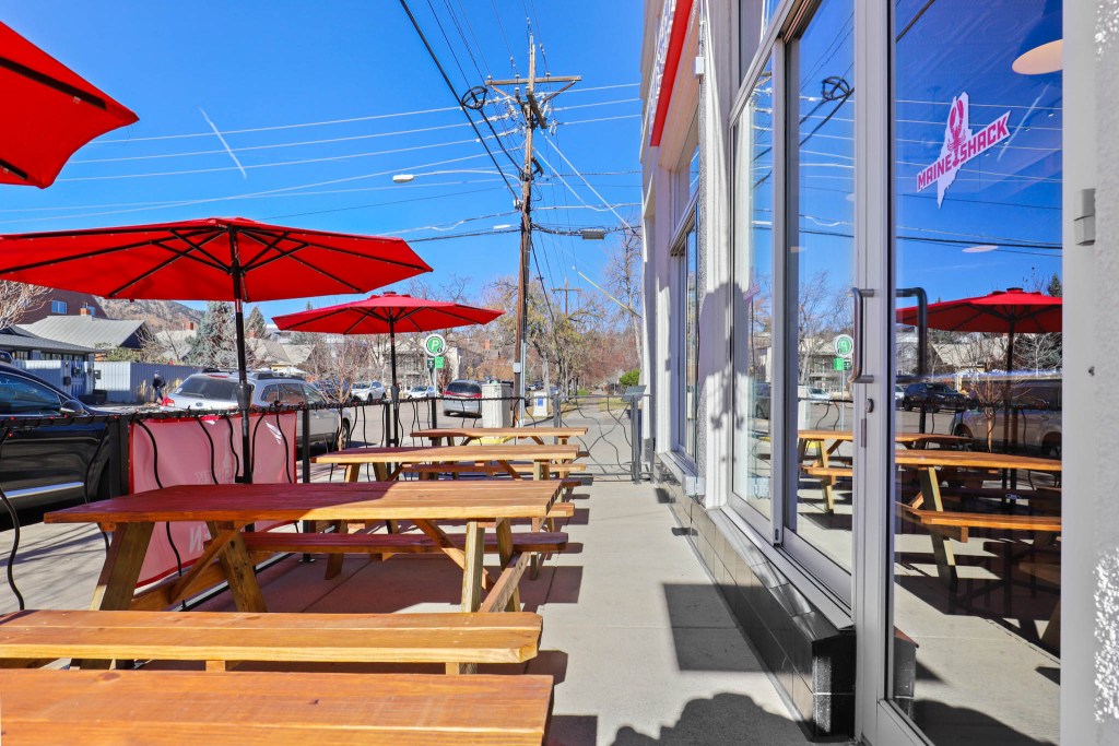 Outdoor seating area of Maine Shack restaurant in Boulder, CO, featuring wooden tables and red umbrellas against a clear blue sky.