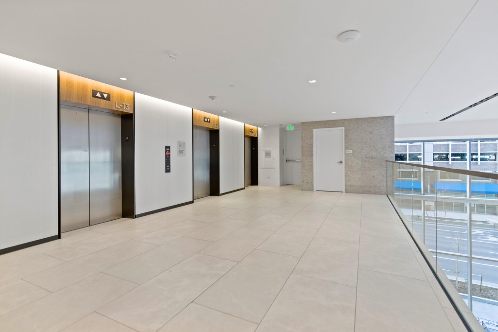 Interior view of a commercial building lobby showing several elevators with illuminated floor indicators, a sleek tiled floor, and modern design elements.
