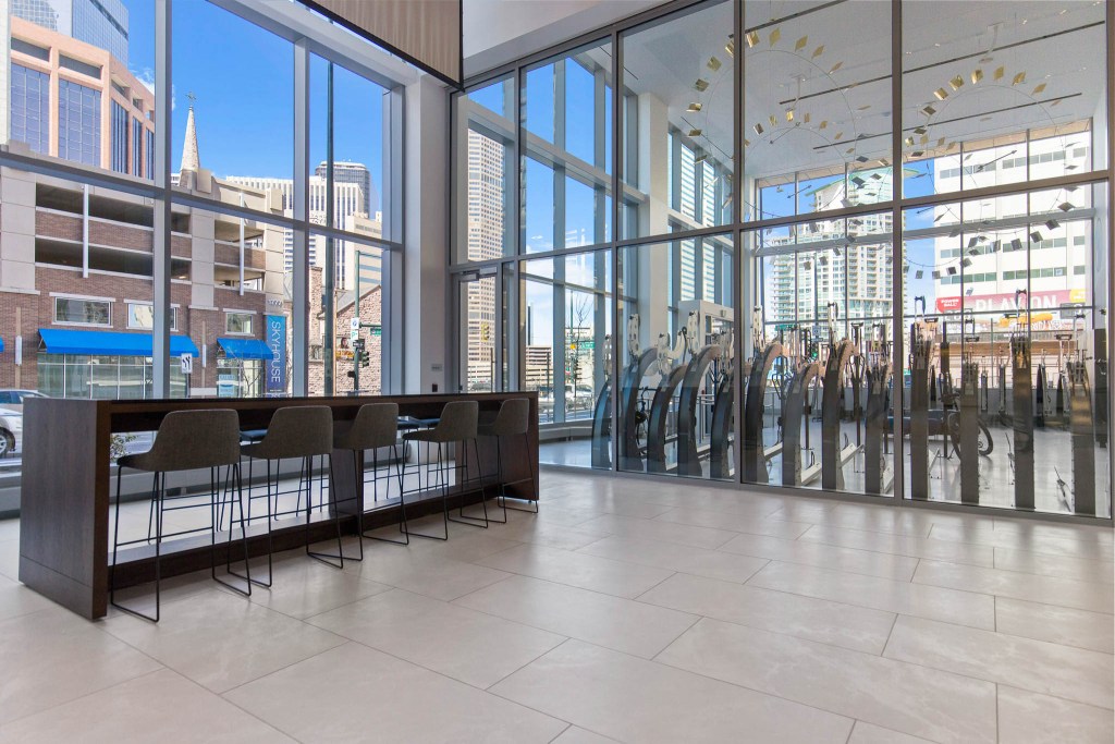 Interior view of a modern commercial space featuring a seating area with high chairs and a bar table, large glass windows showcasing the cityscape outside, and fitness equipment visible through the glass.
