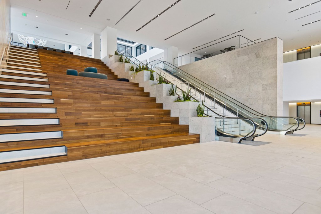 Interior view of a commercial space featuring a wooden staircase with white accents, green plants, and glass escalators.