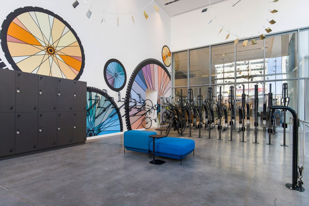 Interior view of a modern bike storage area featuring a colorful mural of bicycle gears, metal lockers, and a blue seating area.