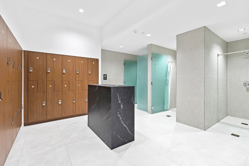 Interior view of a modern locker room featuring wooden lockers, a sleek black countertop, and glass shower stalls with tiled walls.