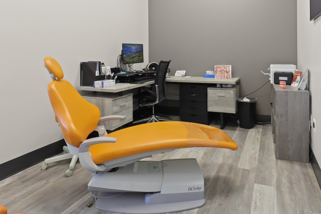 Interior of a dental office featuring an orange dental chair, a workstation with a computer, and a clinical setup.