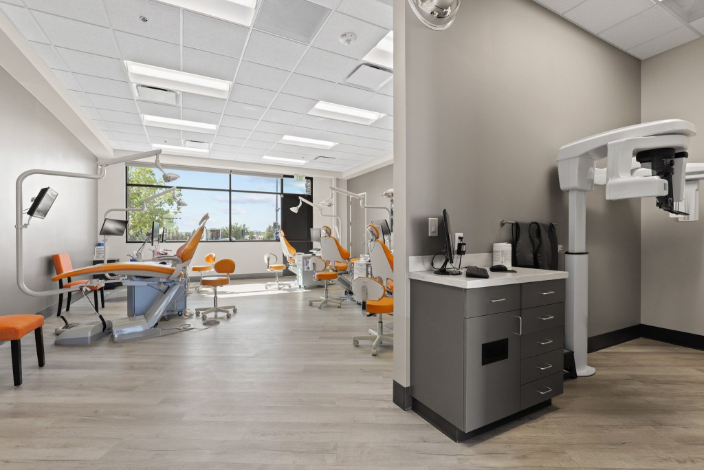 Interior view of a modern dental office featuring orange chairs, treatment chairs, and dental equipment, with large windows providing natural light.