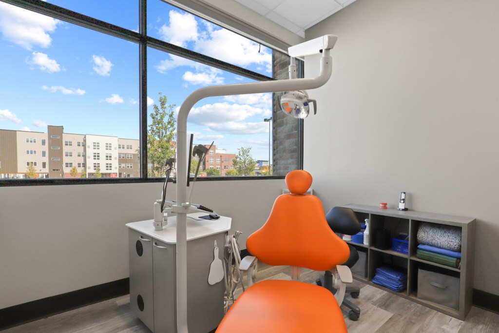 Interior view of a dental office featuring an orange dental chair, dental equipment, and large windows with a view of the outside.
