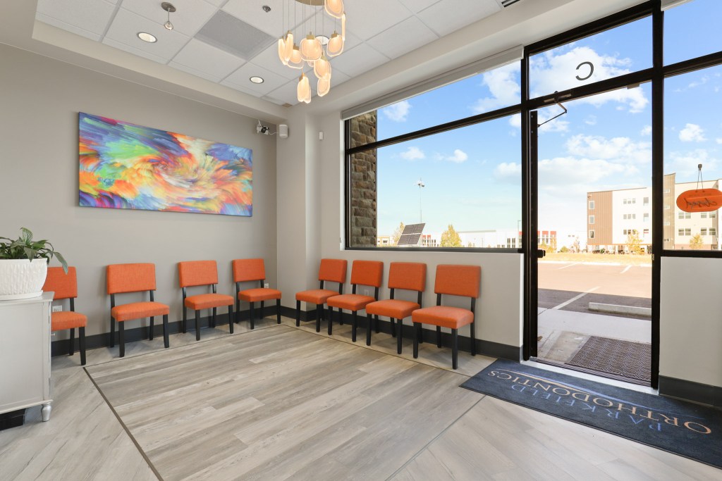 Modern dental clinic waiting area featuring orange chairs, abstract art on the wall, and large windows with natural light.