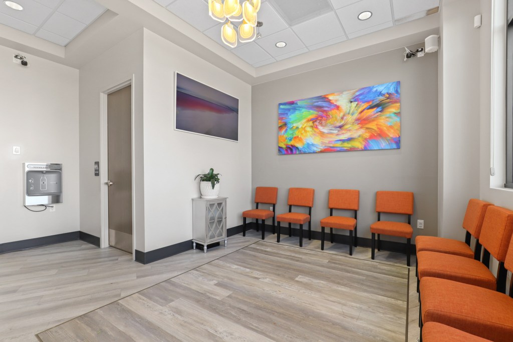 Modern waiting area in a dental or medical facility featuring orange chairs, wooden flooring, a potted plant, and colorful artwork on the wall.