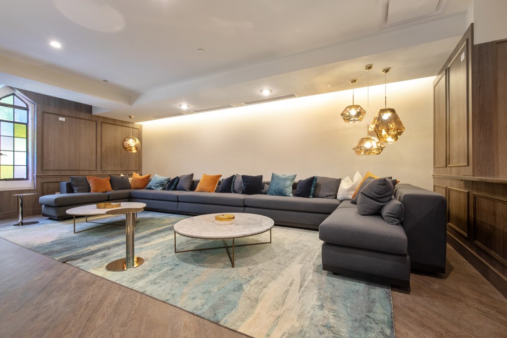 Modern living room featuring a large gray sectional sofa with colorful pillows, a round marble coffee table, and stylish pendant lights, set against a backdrop of wood paneling and a stained glass window.