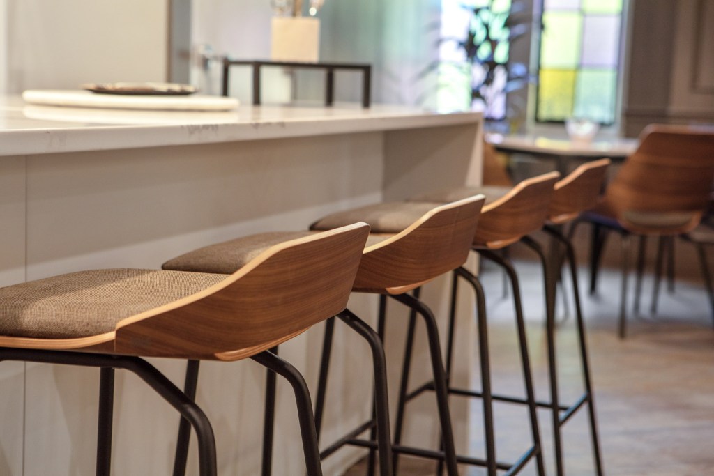 Close-up of modern bar stools with wooden backs and fabric seats lined up at a kitchen island, with a blurred interior background.