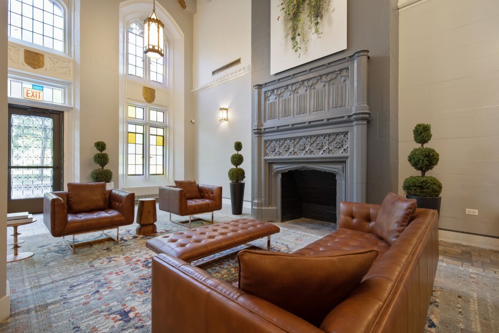 Interior view of a stylish lounge area featuring brown leather sofas, a coffee table, a decorative fireplace, and greenery, with stained glass windows in the background.
