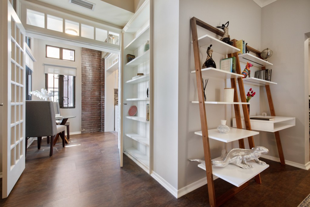 Interior view of an apartment showcasing a hallway with wooden flooring, a decorative ladder shelf with various items, and a dining area with gray chairs and a brick wall in the background.