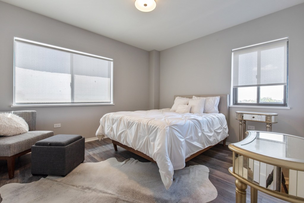 A well-lit bedroom featuring a queen-sized bed with white bedding, a cozy chair, a small ottoman, a decorative rug, and a window with sheer blinds.
