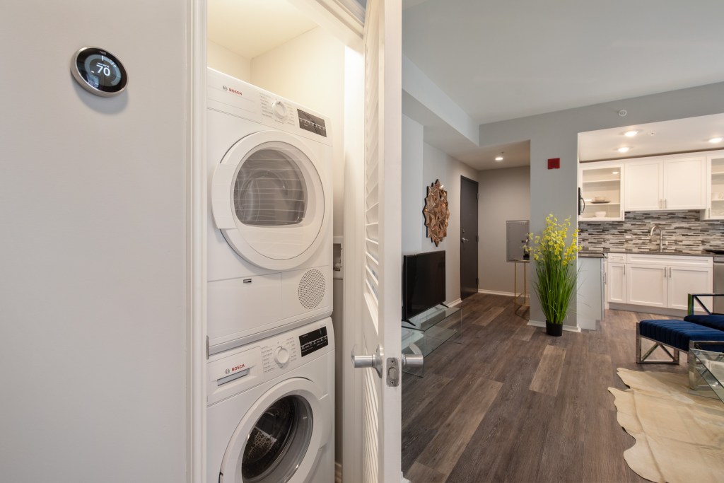 View of a modern apartment interior featuring a stacked washer and dryer, a thermostat displaying the temperature, and an open floor plan with a kitchen and living area in the background.