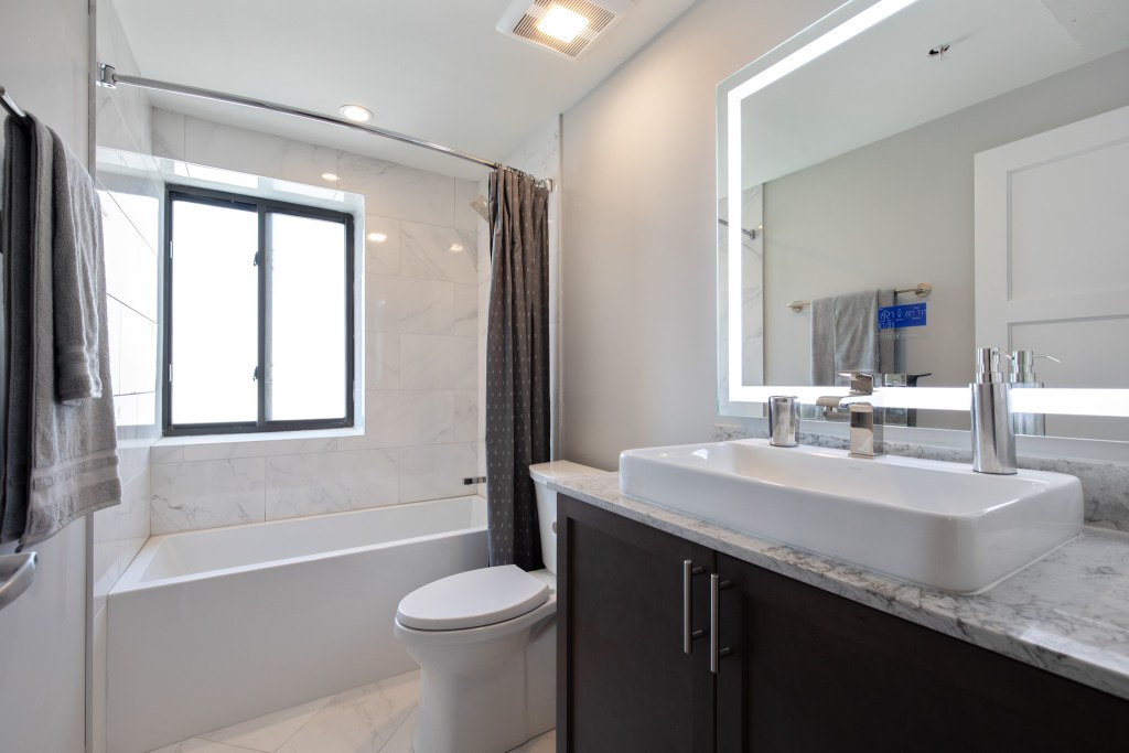 Modern bathroom featuring a white bathtub, a large mirror with integrated lighting, and a stylish sink on a marble countertop.
