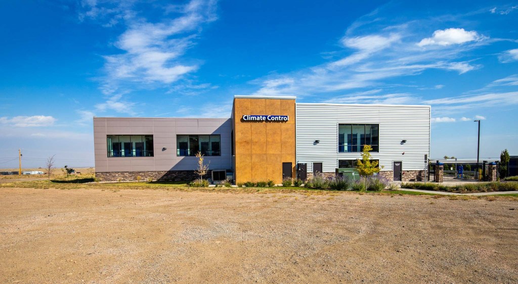 Exterior view of a climate-controlled storage facility with a modern design, featuring large windows and a landscaped entrance under a clear blue sky.