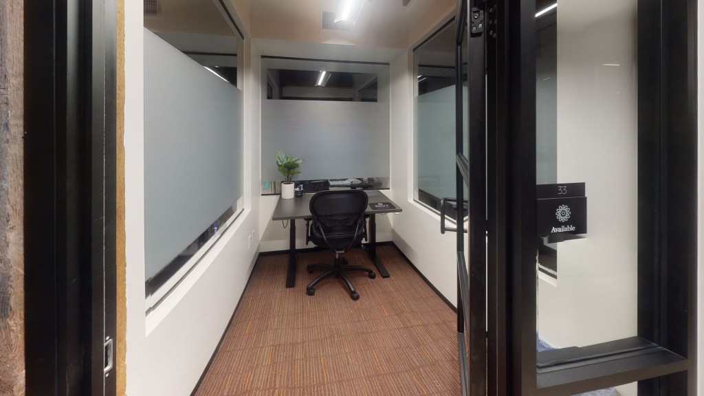 Interior view of a co-working space office, featuring a desk with a chair, a small plant on the desk, and glass partitions allowing natural light, labeled 'Available'.