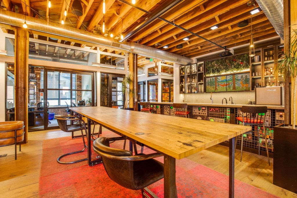 Interior view of a spacious co-working space featuring a long wooden table, leather chairs, large windows, and decorative elements on the walls.