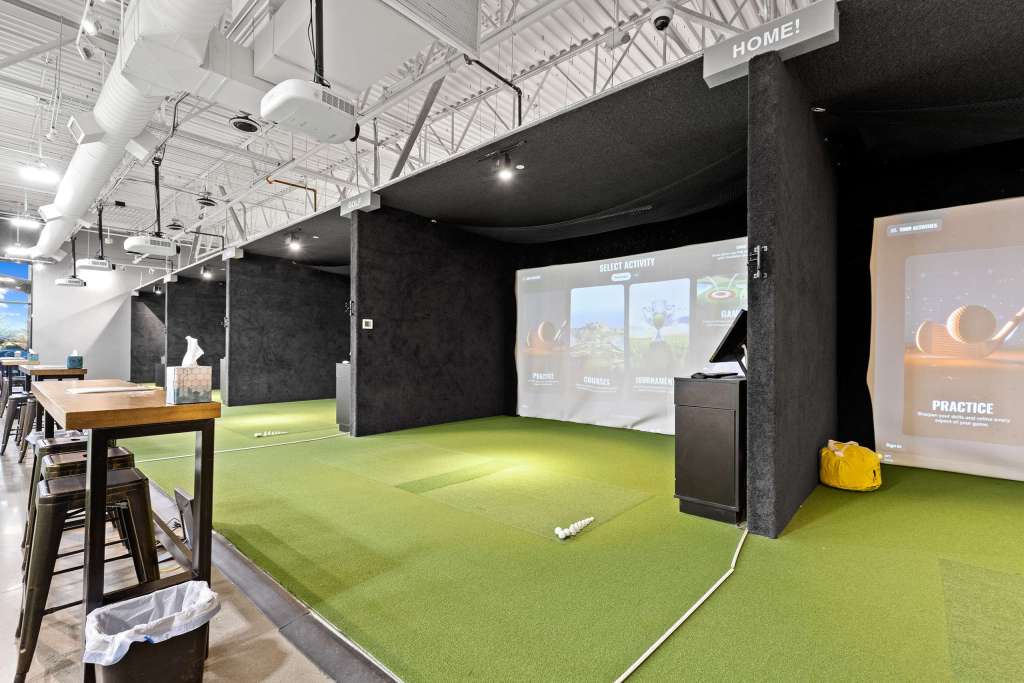 Interior view of The Swing Bays golf simulator with digital screens displaying various activities, green turf flooring, and a seating area with high tables and stools.