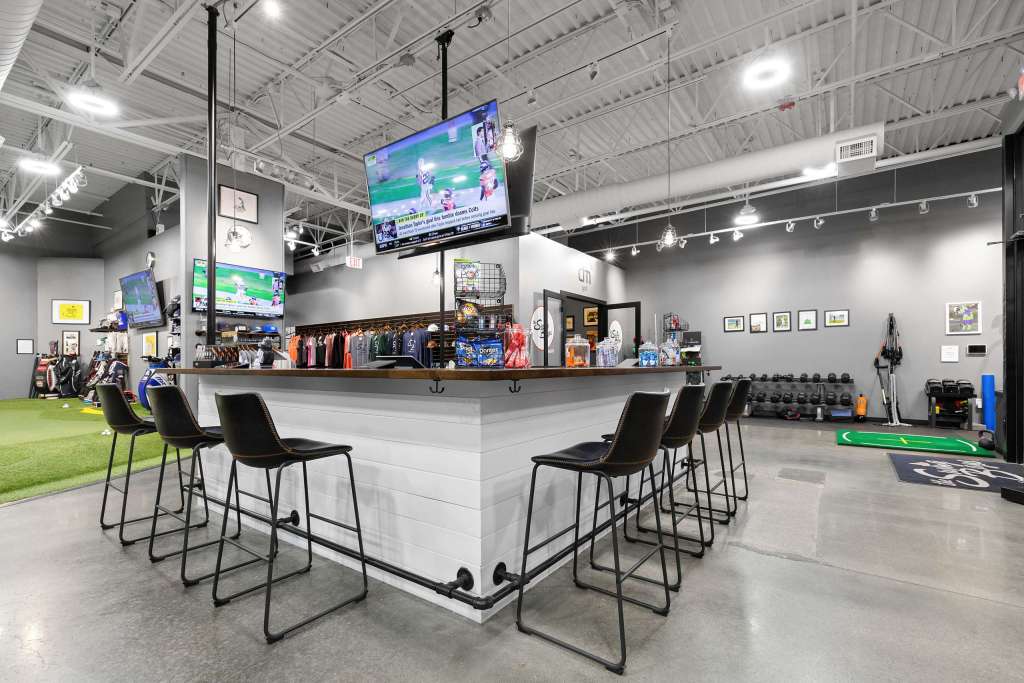 Interior view of The Swing Bays featuring a bar area with high stools, multiple TV screens displaying sports, and a well-organized space for golf simulators.