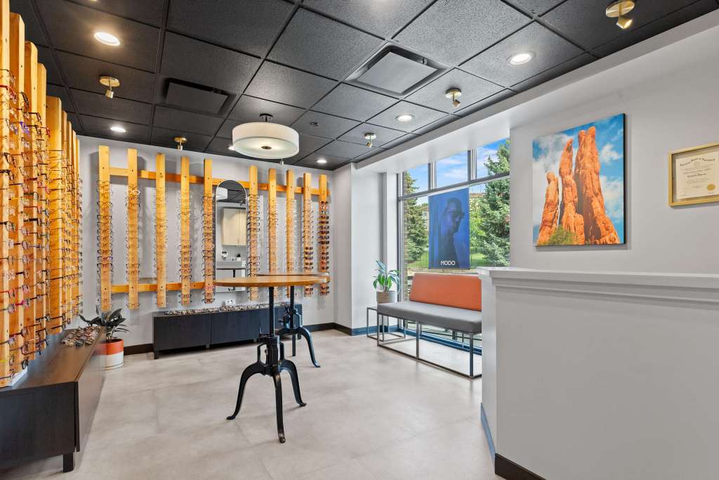 Interior view of an eye clinic showing display racks of eyeglasses on the wall, a waiting area with a bench, and framed artwork. Natural light enters through large windows.