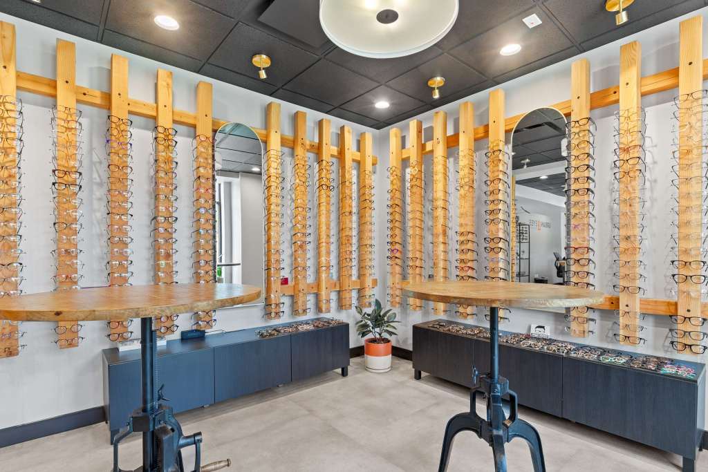 Interior view of an eye care clinic featuring wooden displays of eyeglasses on the walls, a round table in the center, and a mirrored wall behind the display.