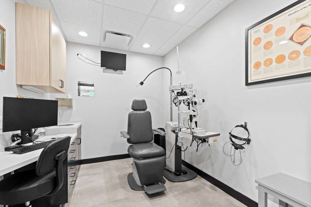 Interior view of an eye examination room featuring a black examination chair, a desk with a computer, and various eye examination equipment.