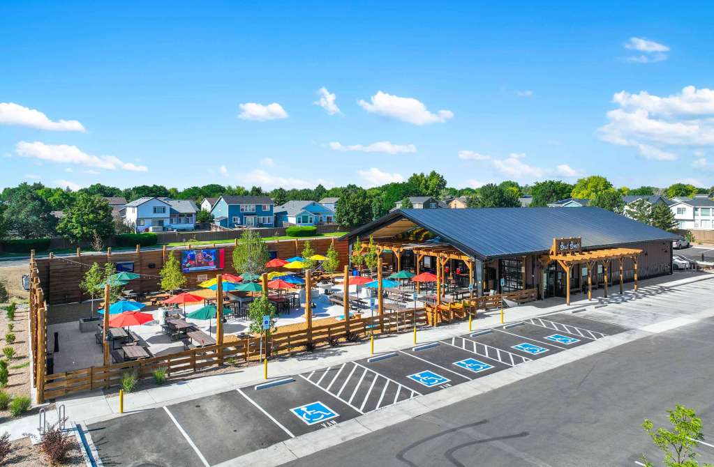 Aerial view of Billy Birch Pub showcasing the expansive outdoor patio with colorful umbrellas, surrounded by greenery and residential buildings under a sunny sky.