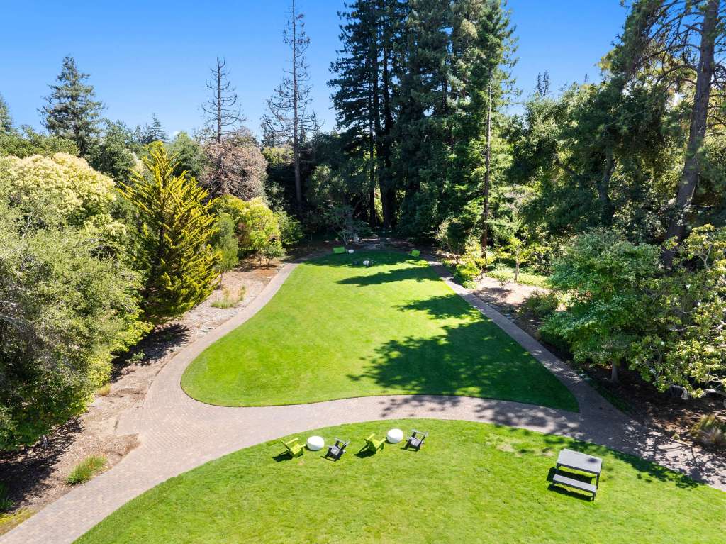 Aerial view of a spacious green lawn surrounded by trees, with outdoor seating including black chairs and white tables, showcasing a natural work setting at Willow Workplace in Menlo Park.